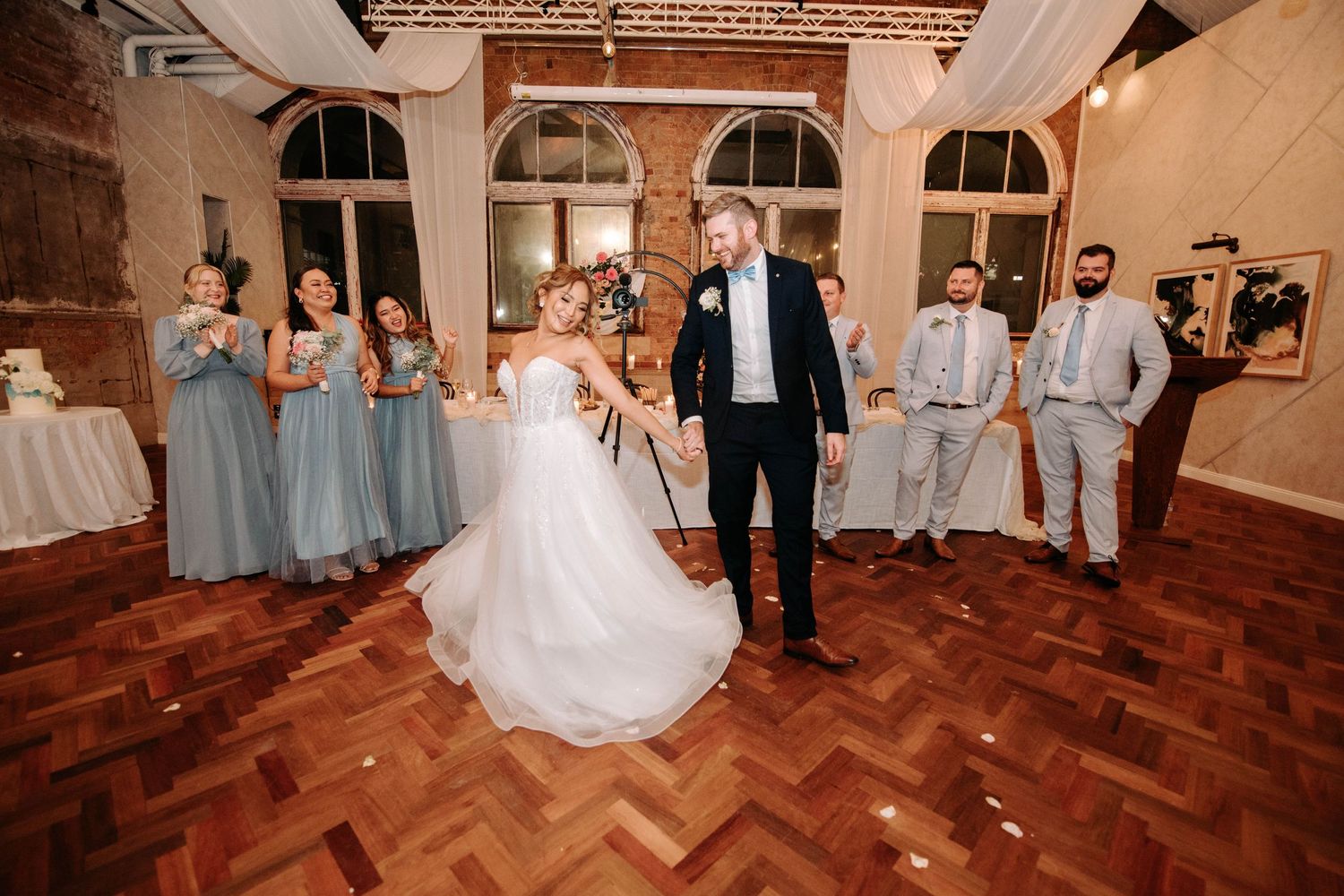 Wedding party dancing on a parquet floor in an elegant brick-walled venue with arched windows.