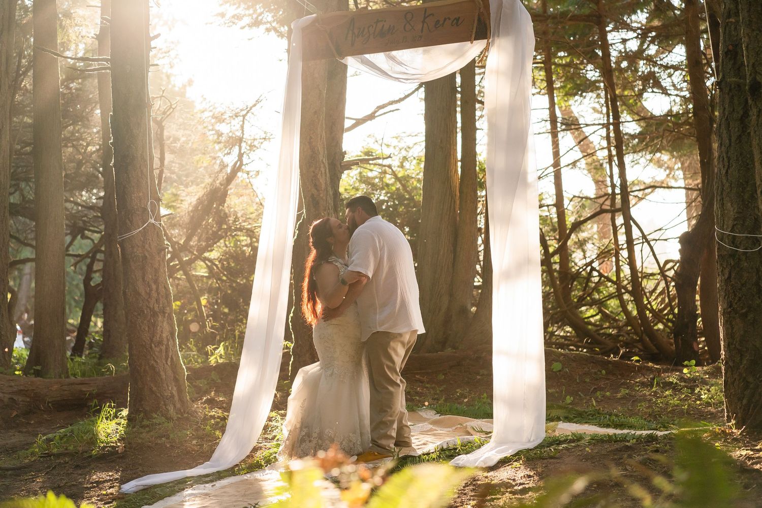 Sunlit wedding ceremony under wooden arch in a forest setting.