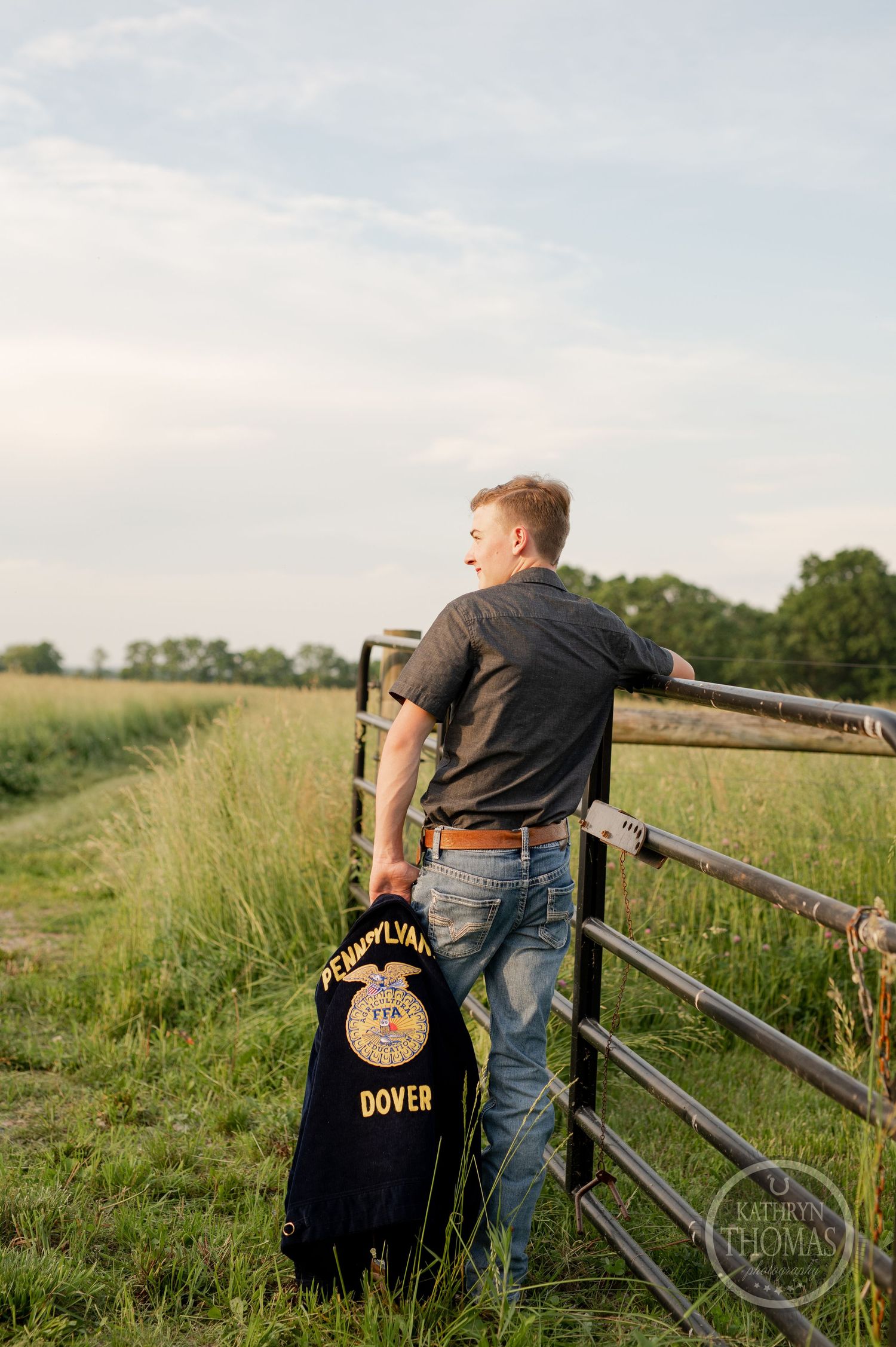 Student holding FFA jacket stands by rustic fence in rural landscape at sunset.