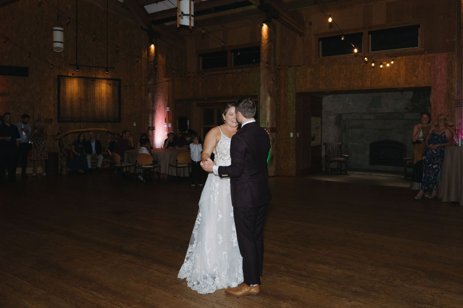 A couple shares their first dance at a rustic wedding reception with wooden floors and ambient lighting.