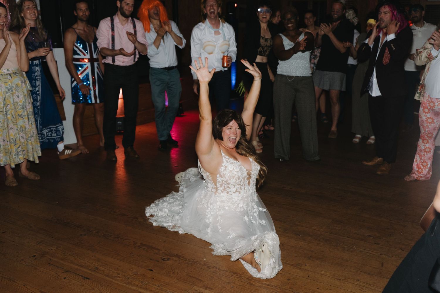 A person in a white wedding dress performs an energetic dance move on a wooden floor surrounded by guests at a reception.