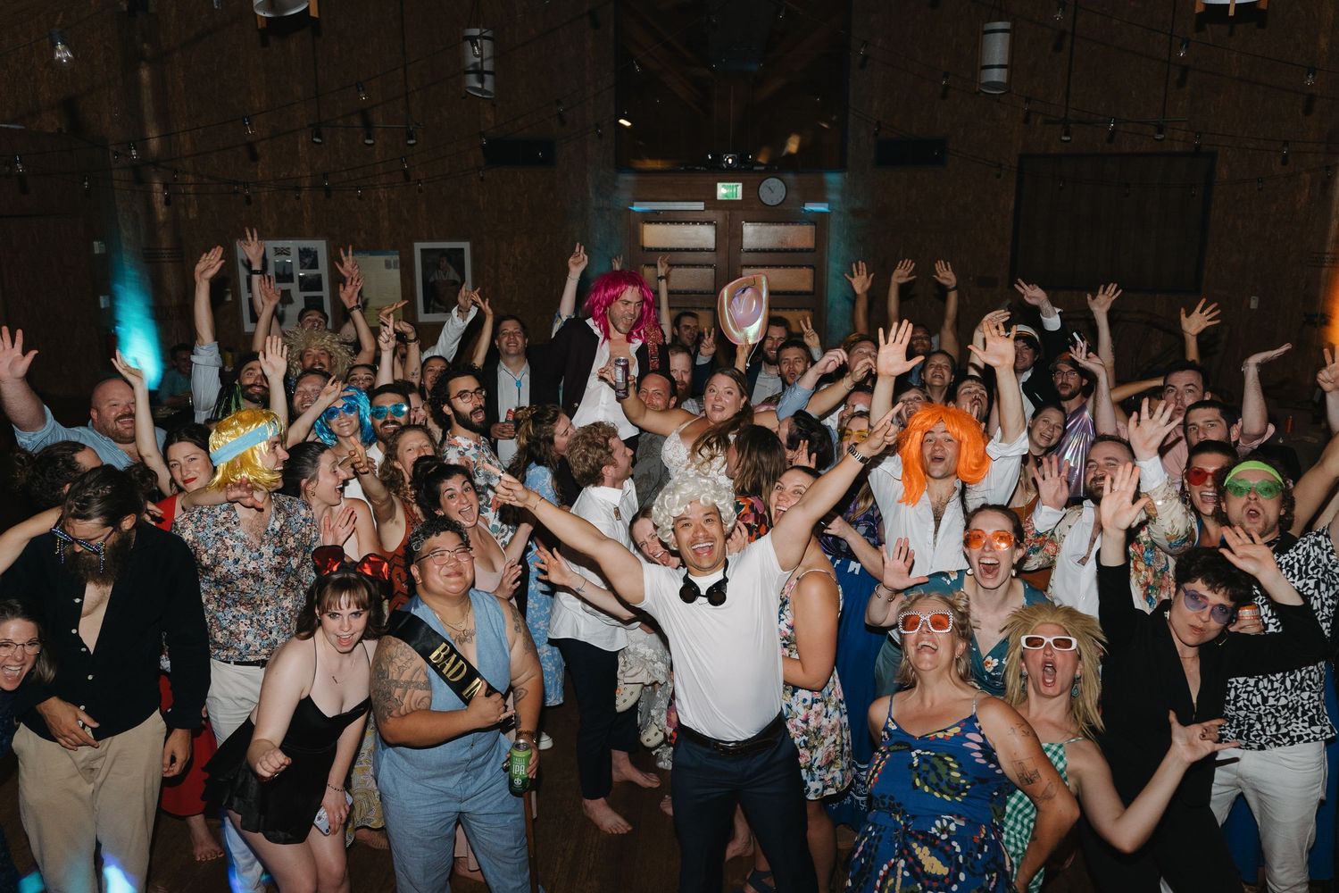 A large crowd of wedding guests dancing and celebrating with their hands in the air on a dark dance floor.