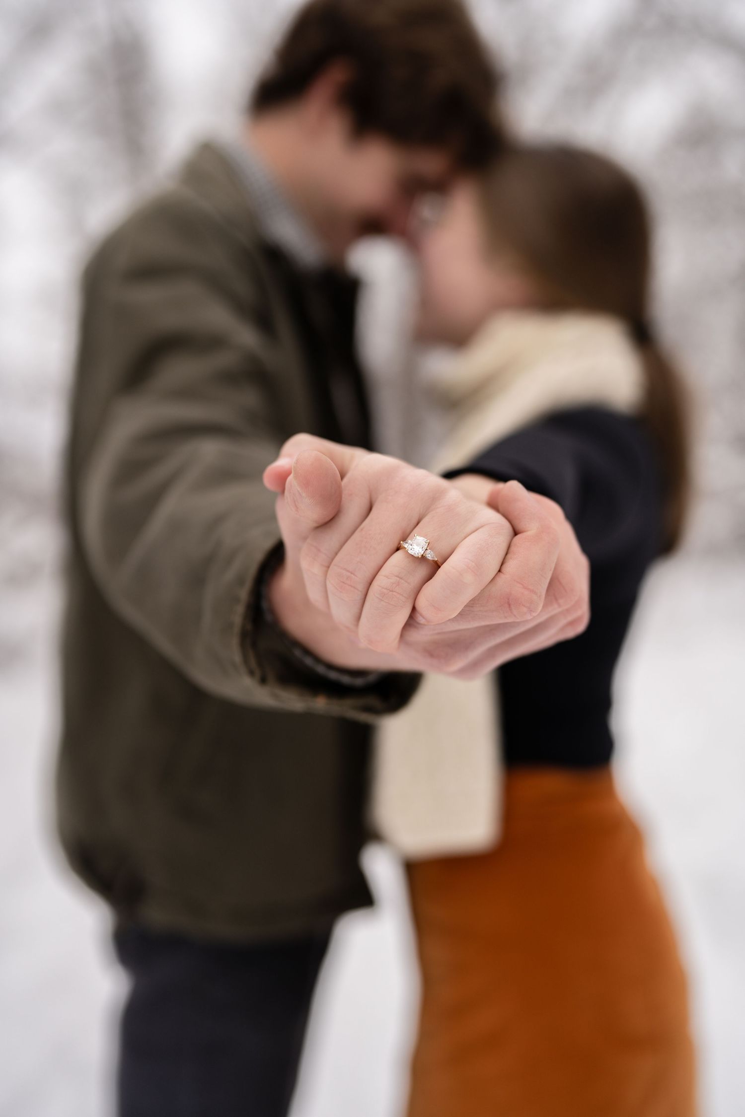 Close-up of an engagement ring on a hand with a romantic couple embracing in the background.