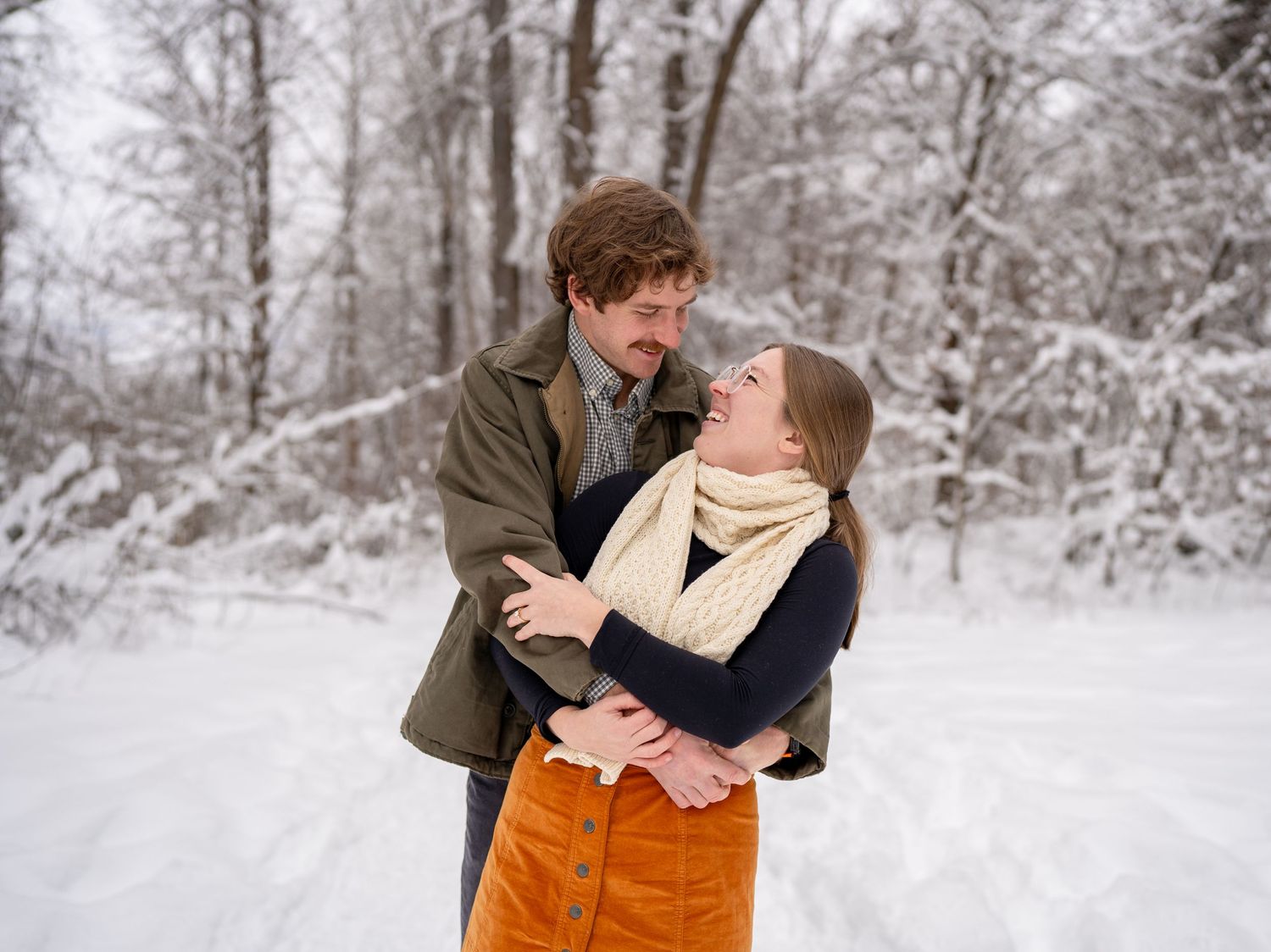 A couple embraces and laughs together in a snowy winter forest setting wearing cozy outfits.
