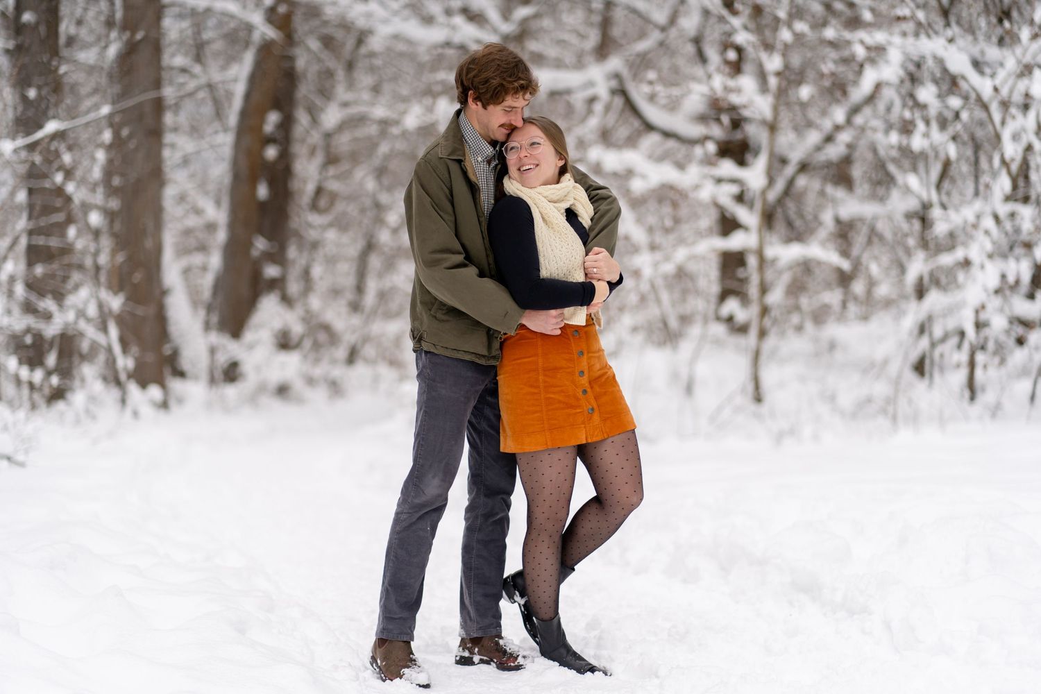 A couple embraces in a snowy winter forest setting with orange skirt adding a bright pop of color against white snow.
