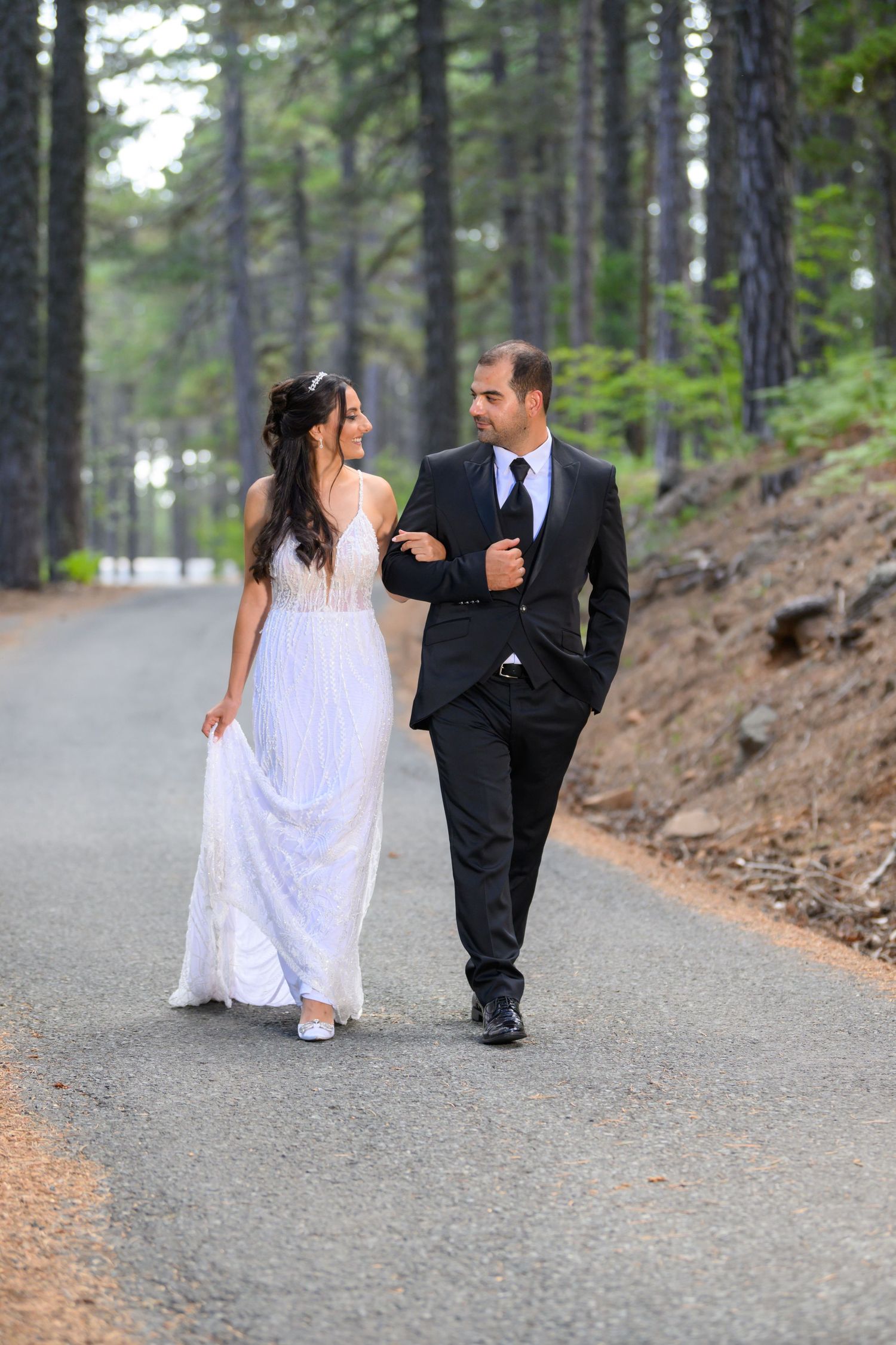A couple walking arm in arm on a forest path, with the bride in a white gown and groom in a dark suit.