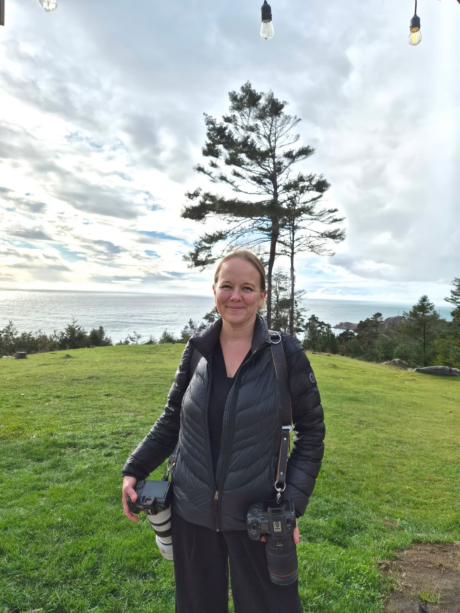 Professional photographer in black dress poses with camera gear at outdoor venue.