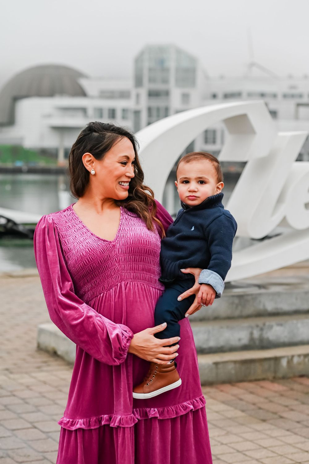 Expectant parent in magenta dress holding toddler against waterfront backdrop.
