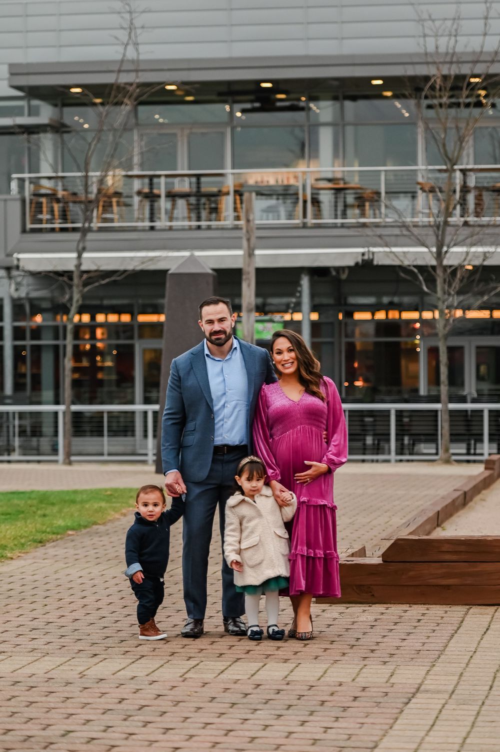 A family of four poses together outside a modern building on brick pavement.