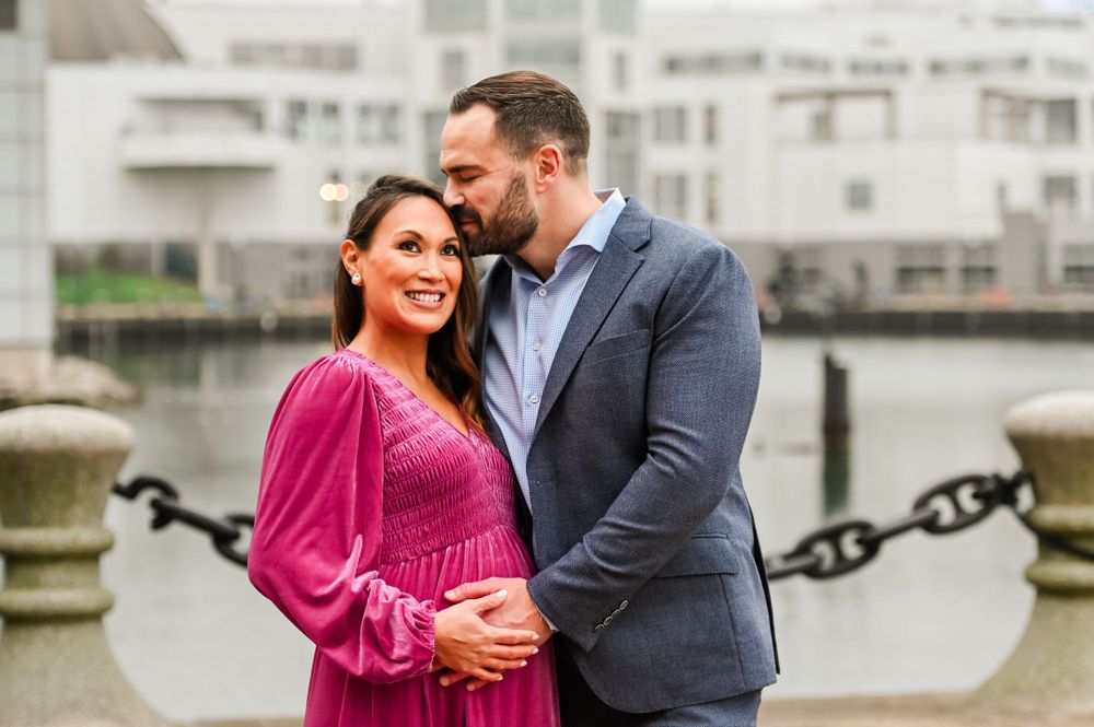 A couple shares an intimate moment by the waterfront with boats and buildings in the background.