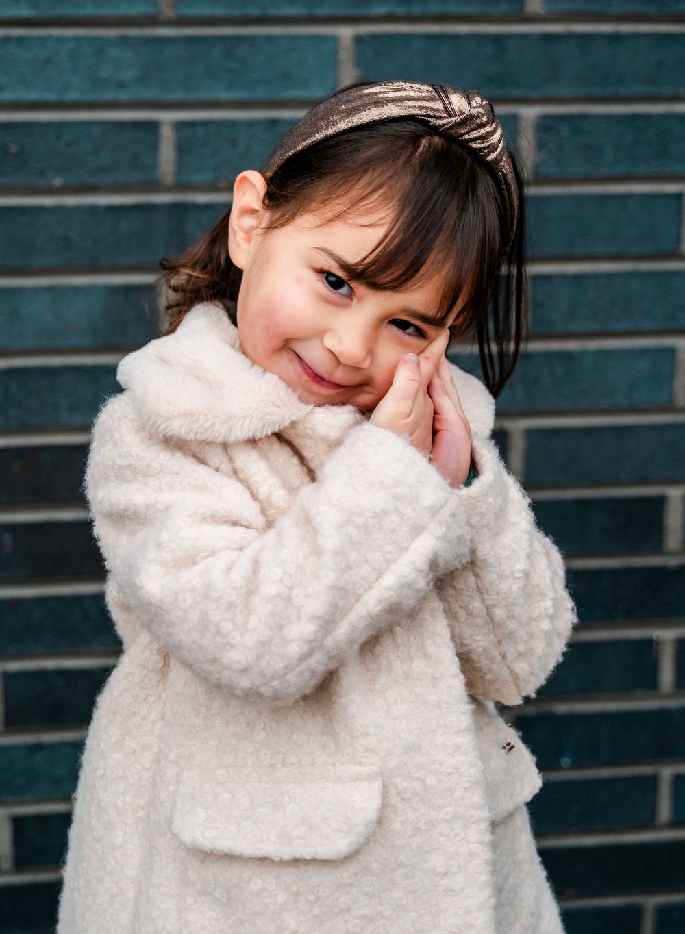 A young child in a cream coat poses adorably against a blue brick wall.