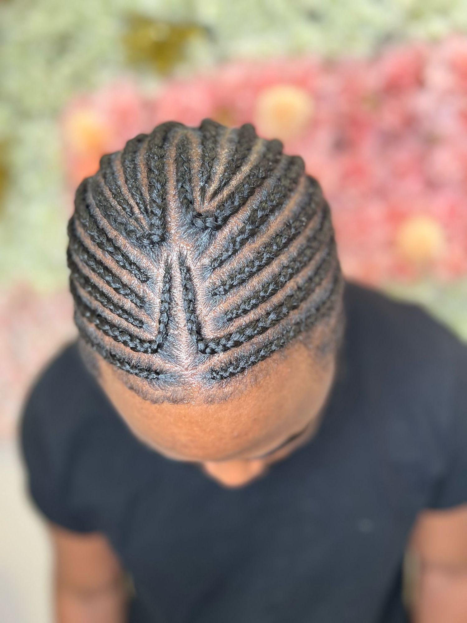Intricate cornrow braiding pattern styled in neat geometric lines on a black background with blurred pink flowers.