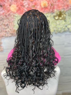 Box braids styled with cascading curls at the ends, shown from behind against a pink and green blurred background.