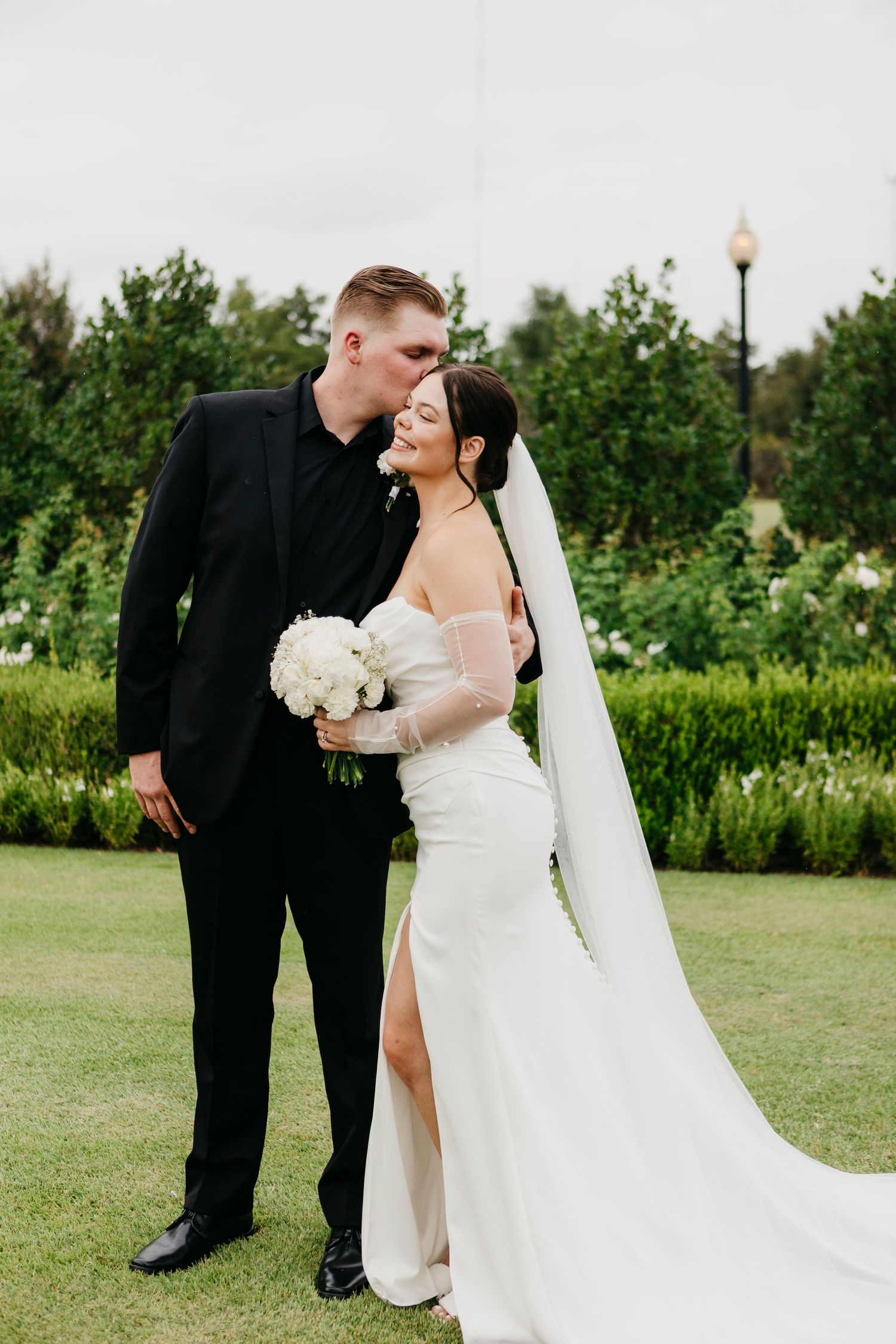 Newlywed couple in formal attire sharing intimate moment on grassy lawn.