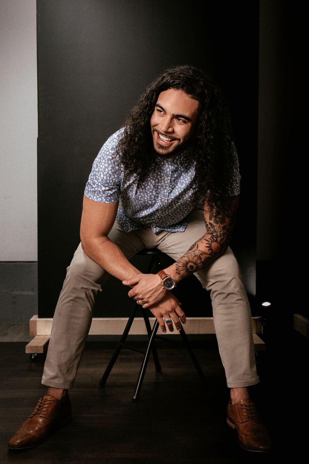 Smiling man with long curly hair and tattoos sits on a stool wearing a patterned shirt and khaki pants.