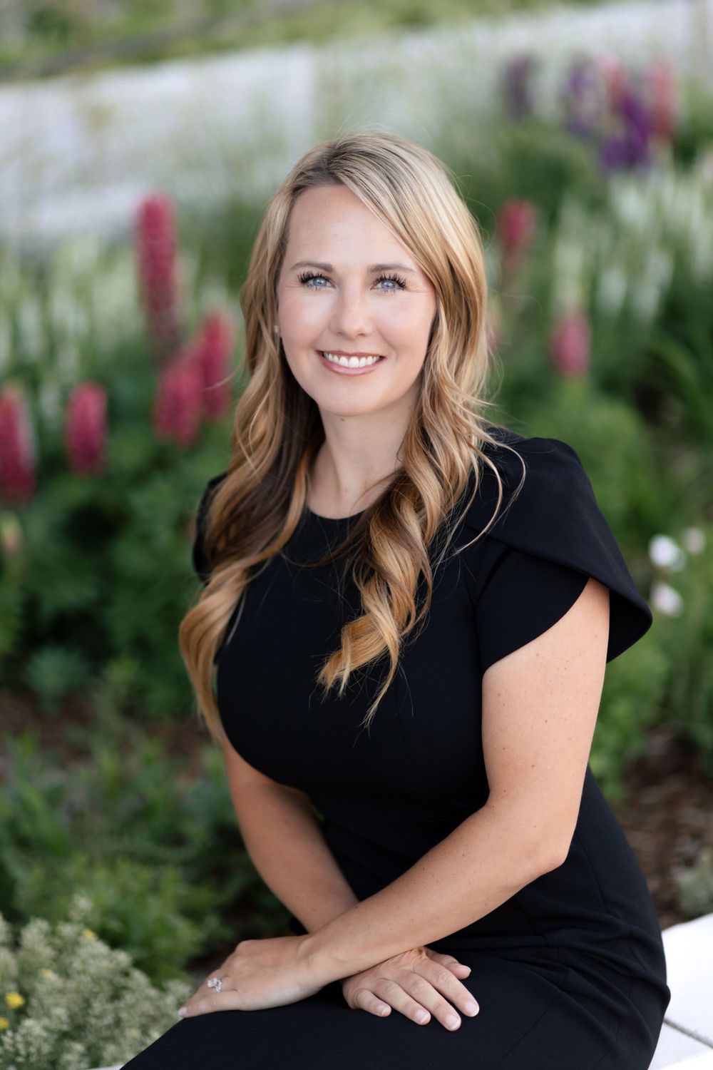 Professional portrait of a woman in a black dress sitting outdoors with colorful flowers in the background.