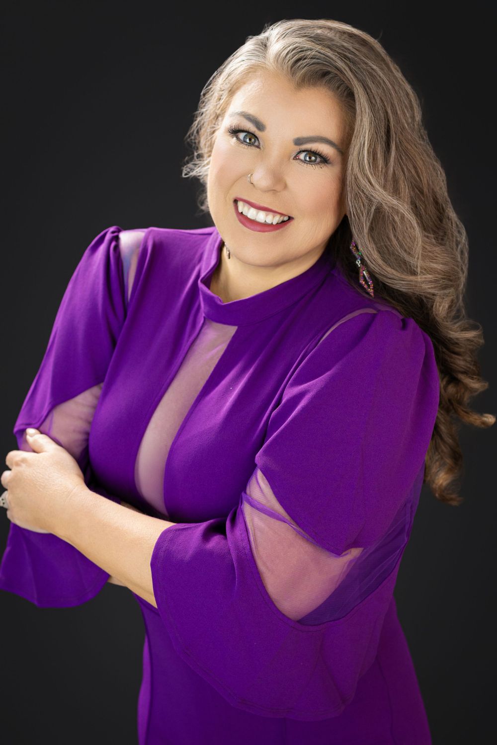A smiling woman in a purple dress with sheer details poses confidently with arms crossed against a dark background.