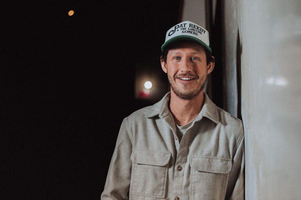 Smiling man wearing a green trucker hat and beige jacket poses against a dark background outdoors.