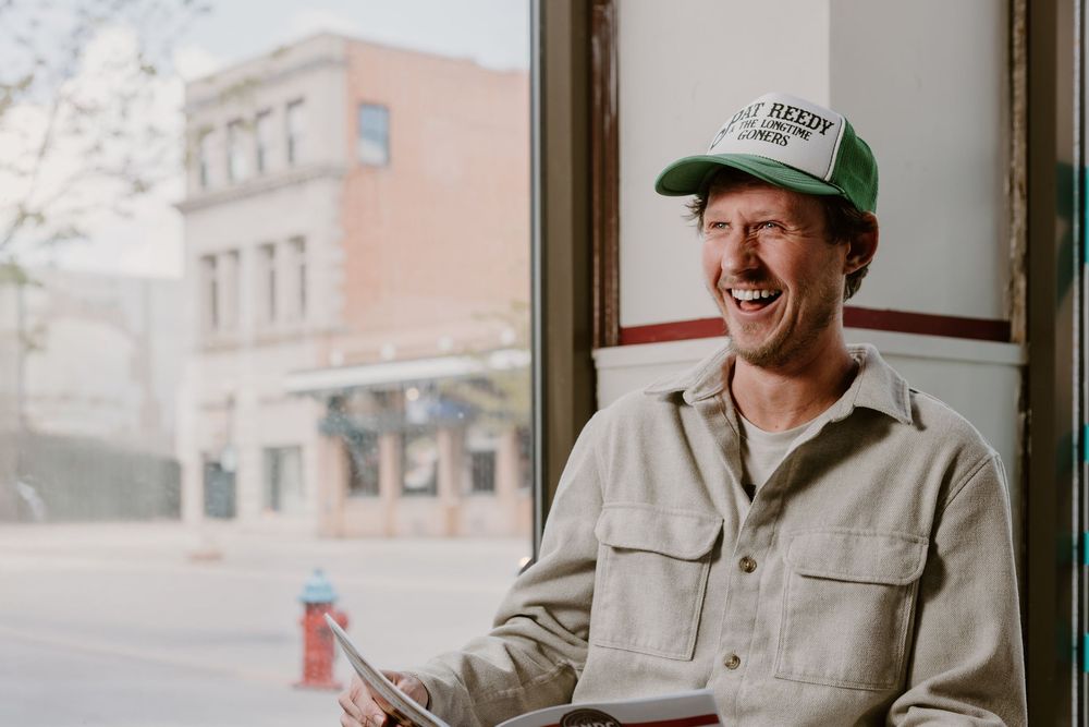 Smiling man wearing a green trucker hat and beige shirt sitting by a window in an urban setting.