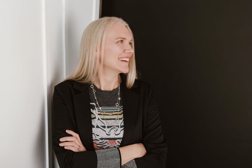 Smiling blonde woman in black blazer and layered necklace standing with arms crossed against a dark wall.