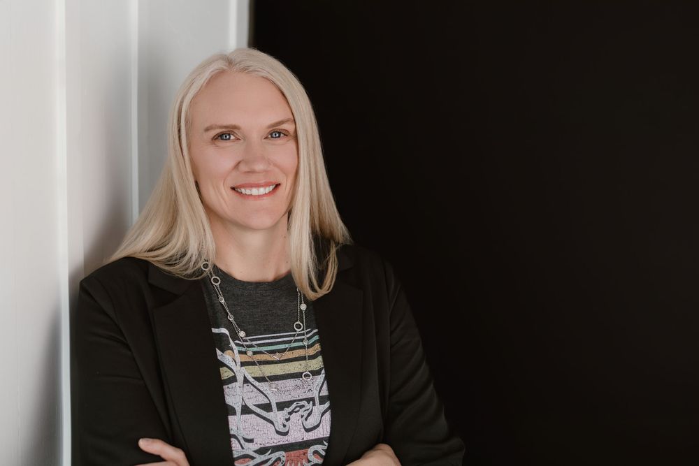 Smiling blonde woman in black sweater with arms crossed, posing confidently against a dark background.