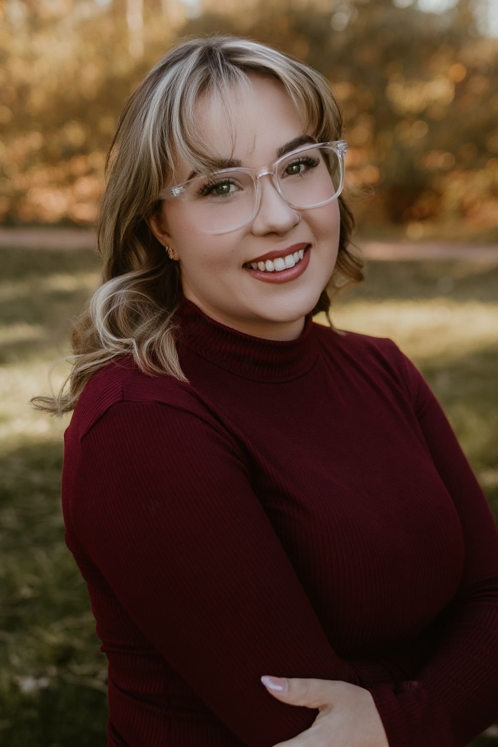 Smiling woman with glasses and blonde hair wearing a burgundy turtleneck sweater, posing outdoors in warm autumn light.