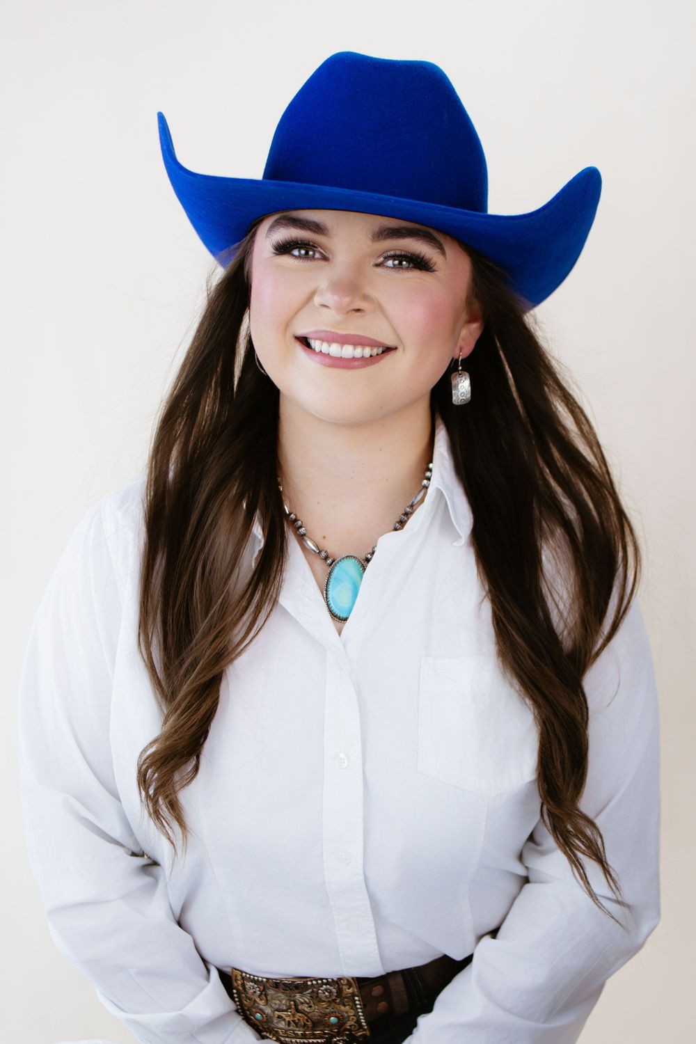 Smiling woman in blue cowboy hat, white shirt, turquoise necklace, and leather belt poses against neutral background.