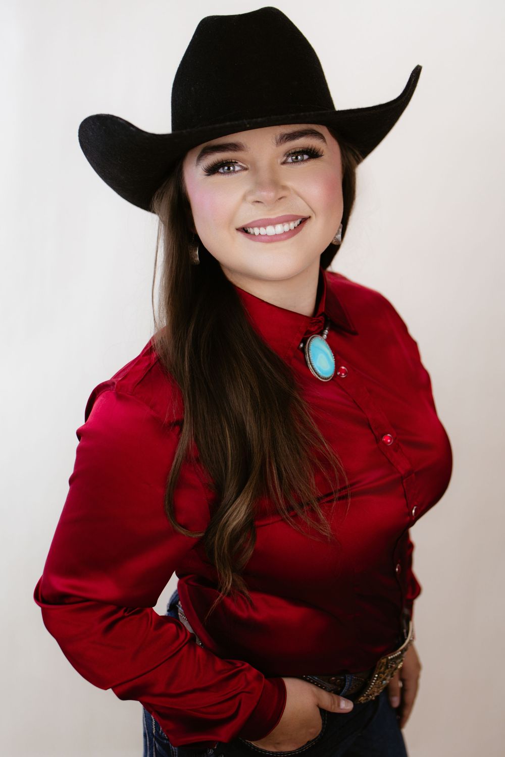 Smiling young woman in black cowboy hat, red western shirt, turquoise bolo tie, and jeans poses against white background.