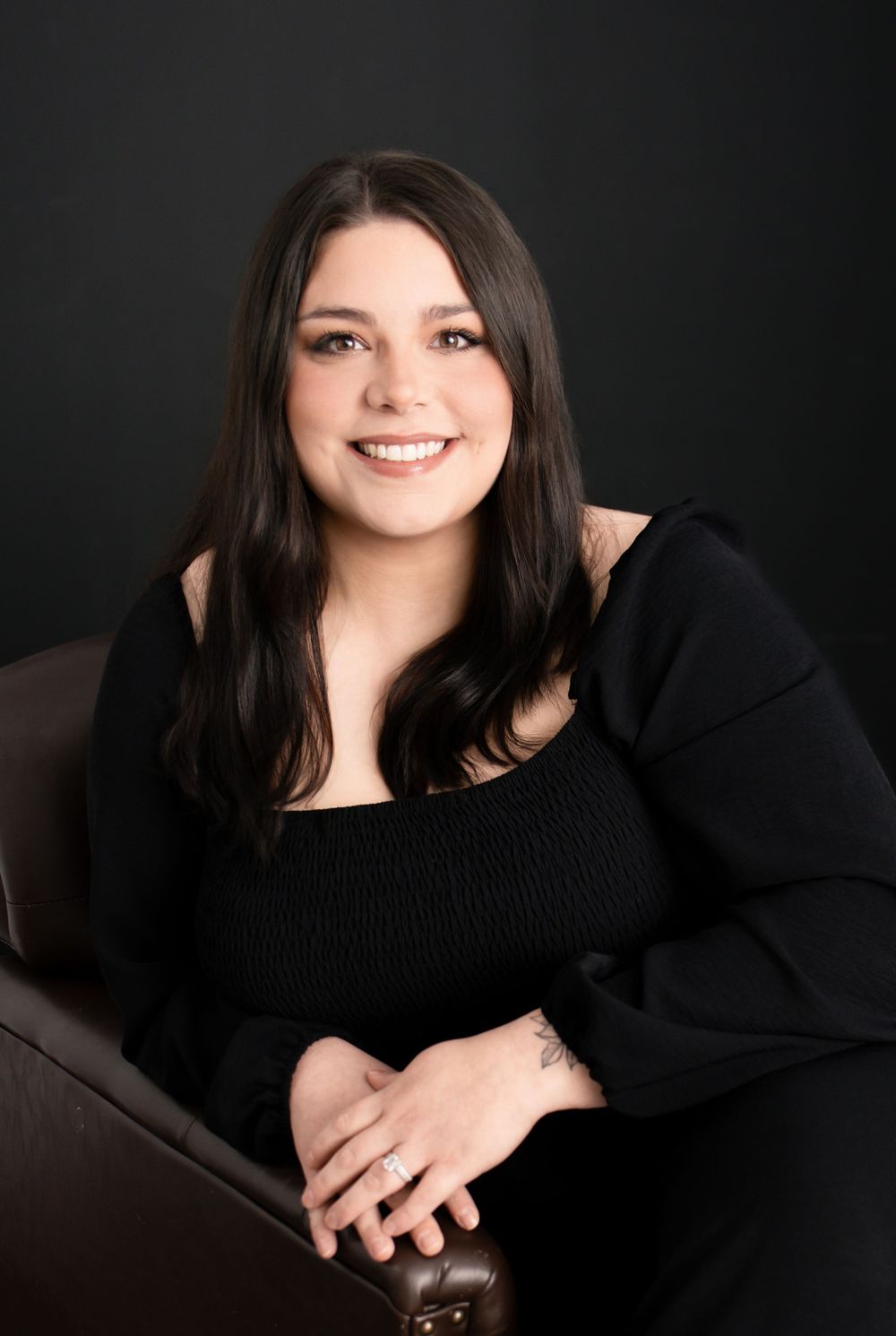 Young woman with dark hair smiling, wearing black off-shoulder top, seated in brown chair against dark background.
