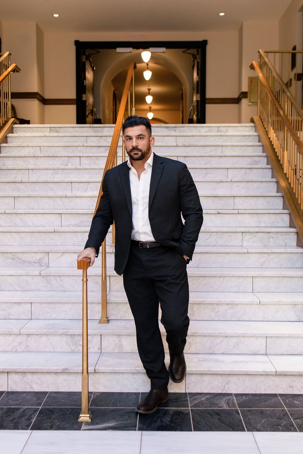 Confident bearded man in black suit descending marble stairs in an elegant building interior.