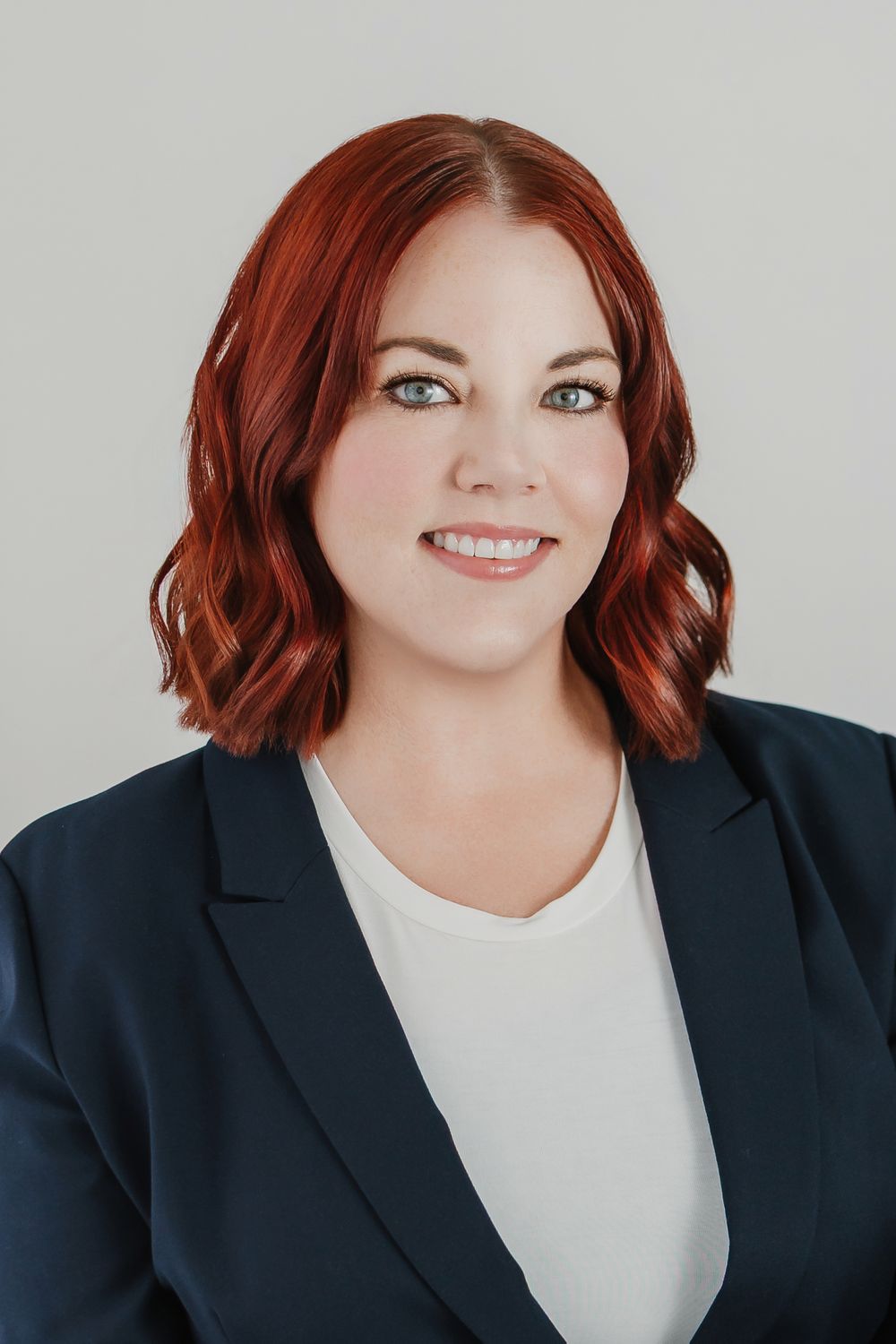 Professional headshot of a smiling woman with red wavy hair wearing a navy blazer over a white top.