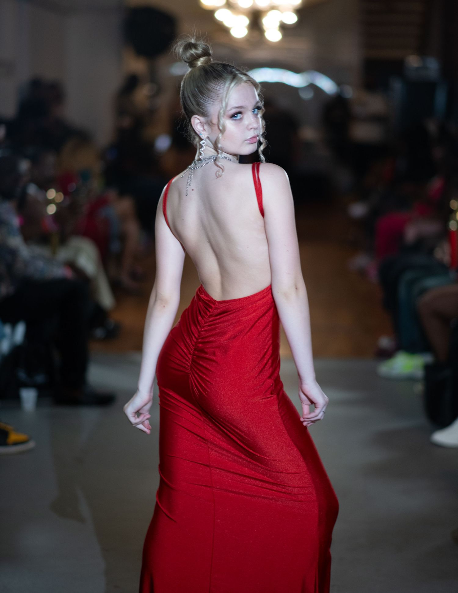 Fashion model in backless red evening gown poses on runway with dramatic lighting.