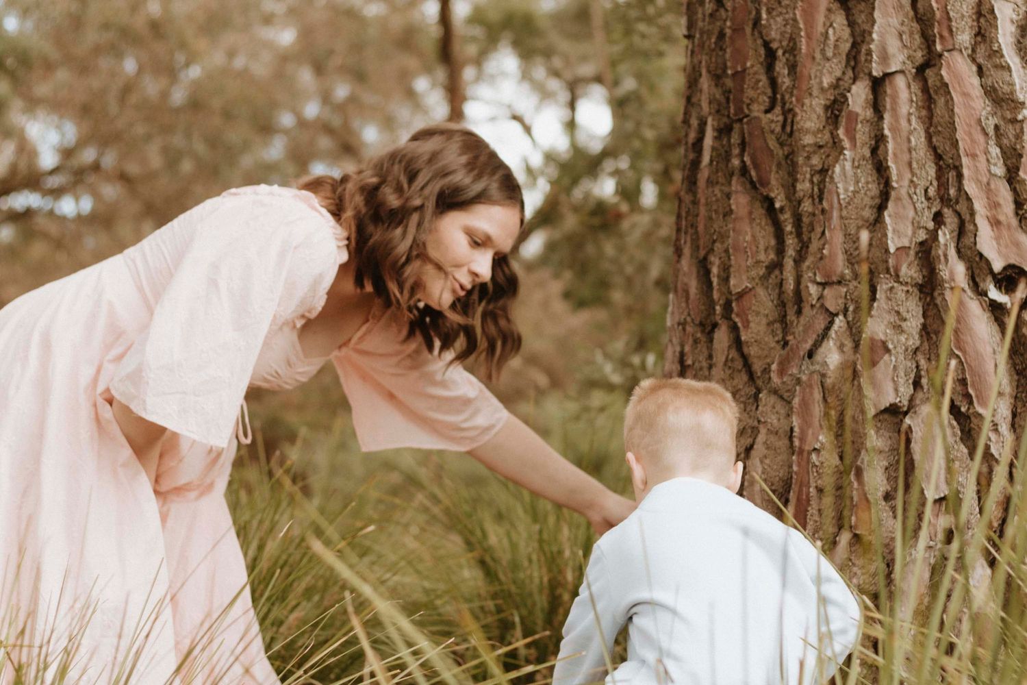A woman in a flowing dress bends down to interact with a small child near a large tree trunk.