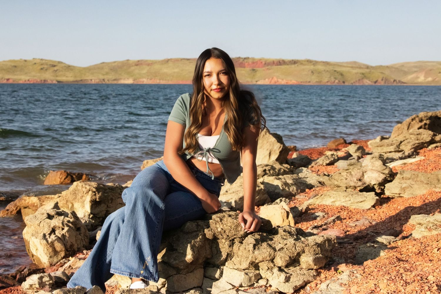 Someone sits on red rocks near a lake with mountains in the background on a sunny day.