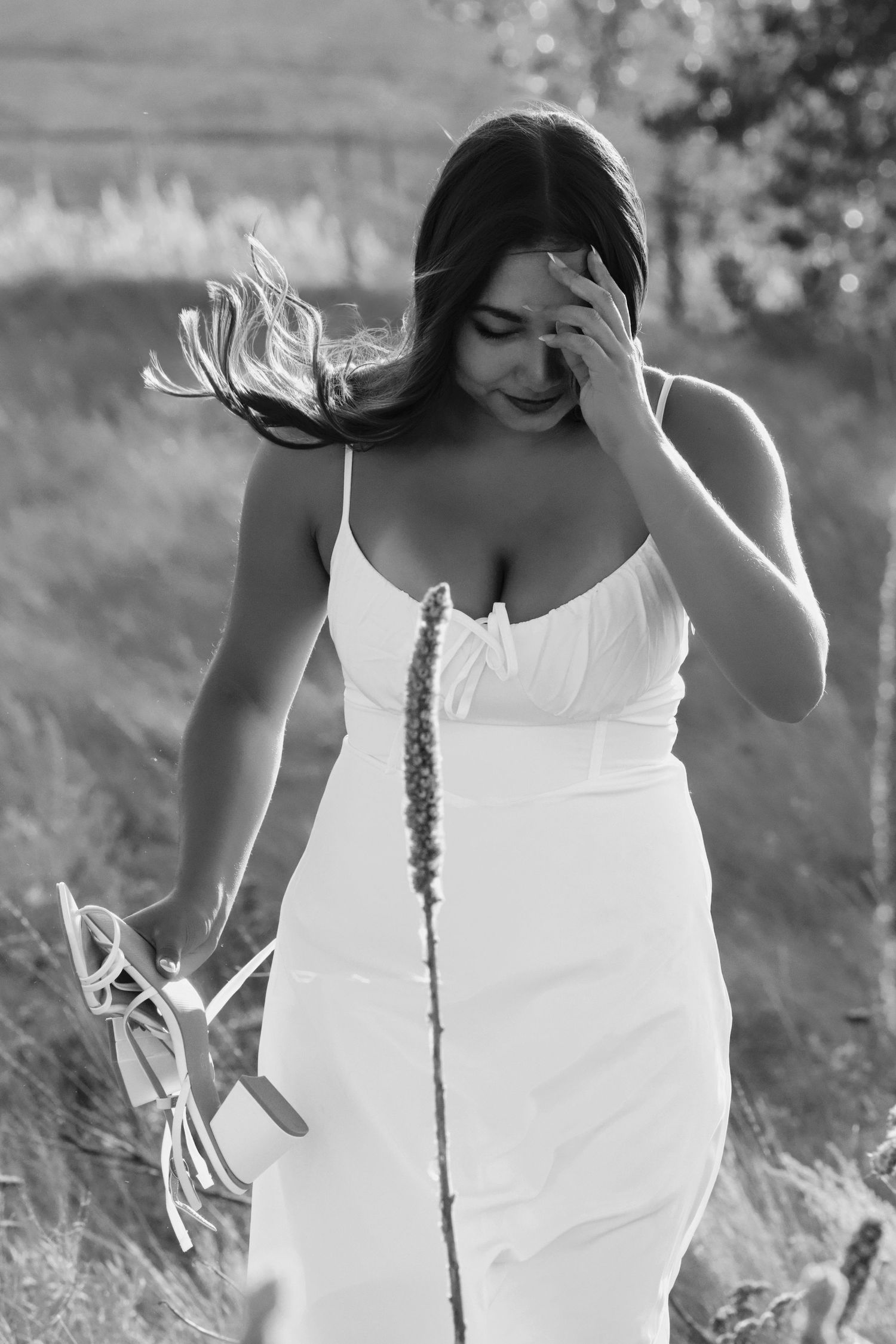 A woman in a white sundress walks through a field holding shoes while her hair blows in the wind.