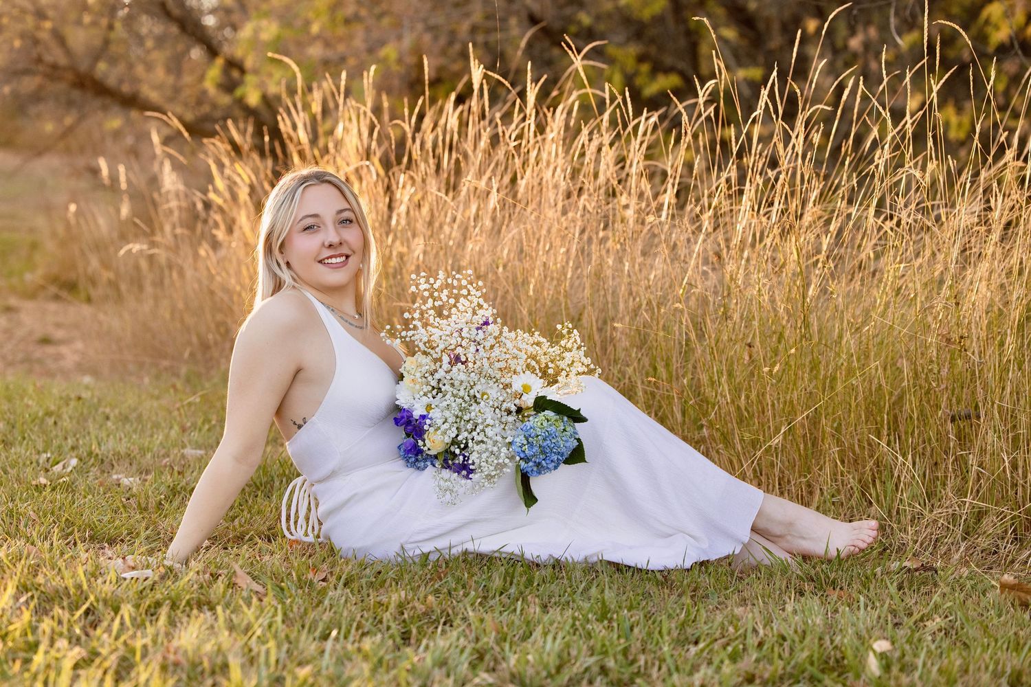 A person in a white dress poses in a golden field during sunset while holding a bouquet of flowers.