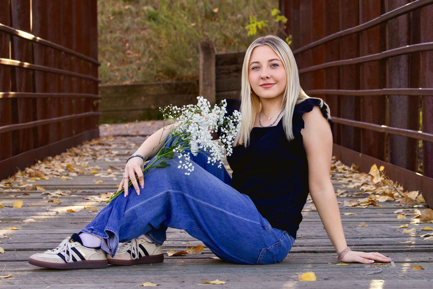Person in black top and blue jeans sitting on wooden bridge with autumn leaves and holding white flowers.