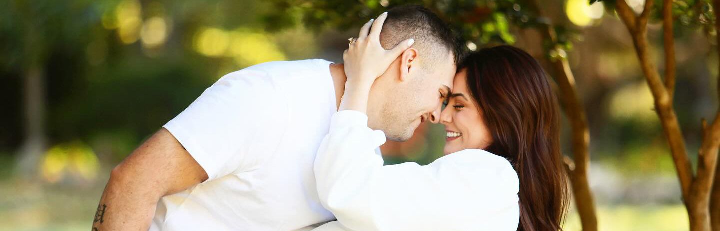 A romantic couple in white shares an intimate moment outdoors in dappled sunlight.