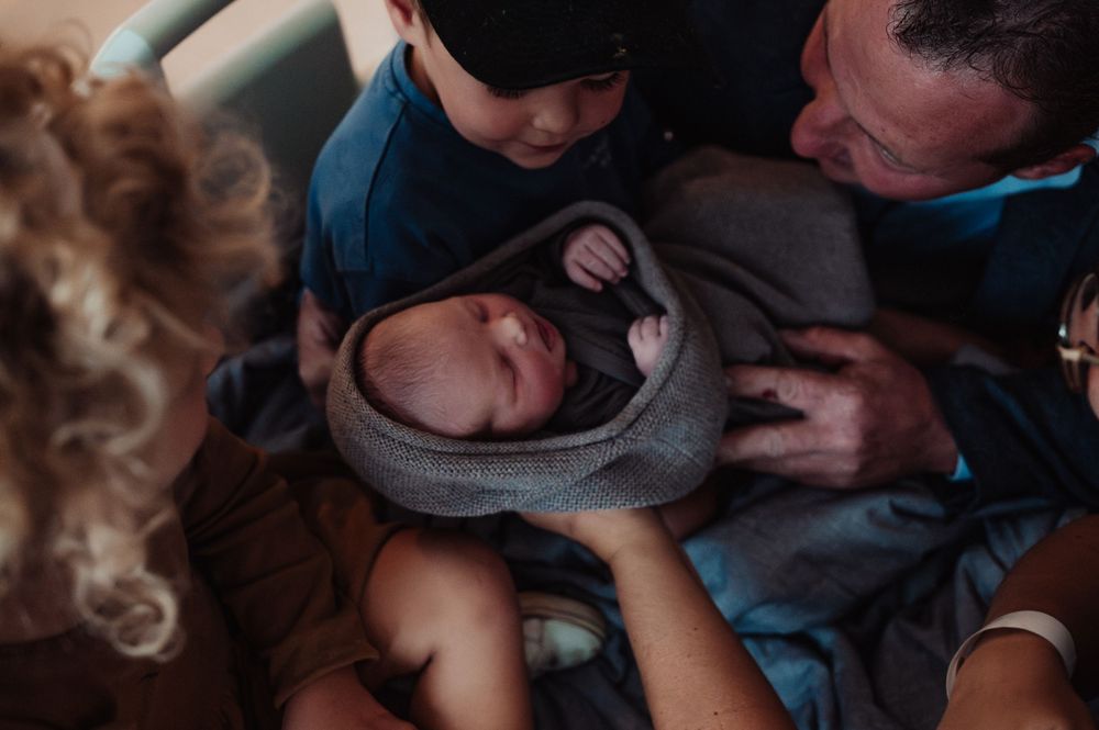 A small newborn wrapped in a gray blanket is surrounded by caring observers in a medical setting.