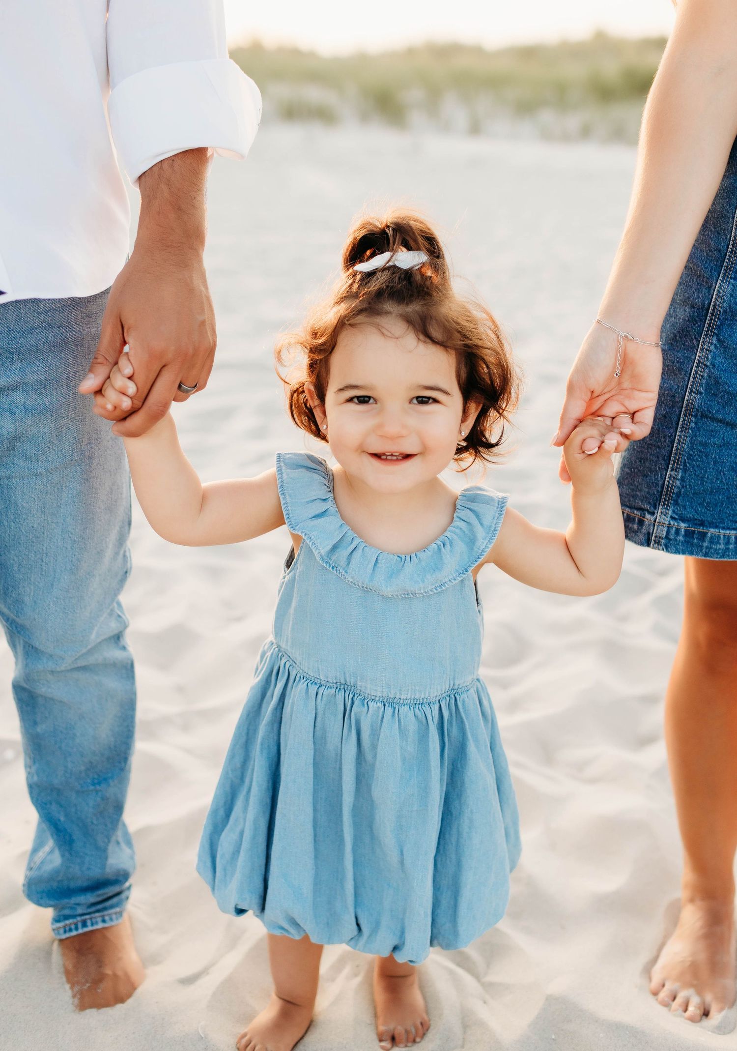 Parent in hat holds child in yellow dress by rustic fence at sunset.