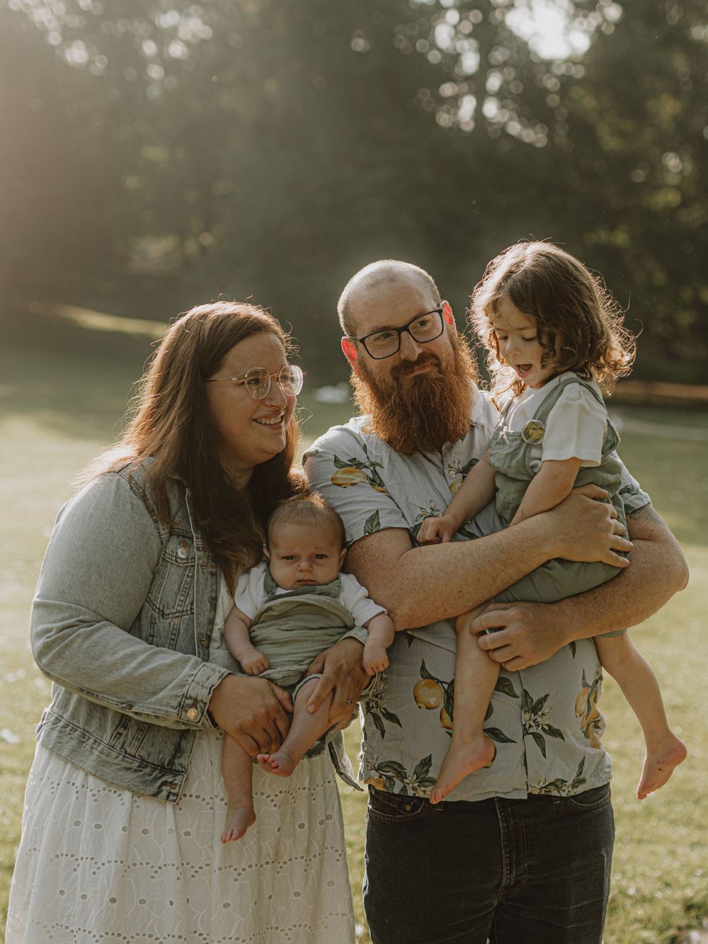 Family portrait in a park with warm sunset lighting showing parents with two young children.