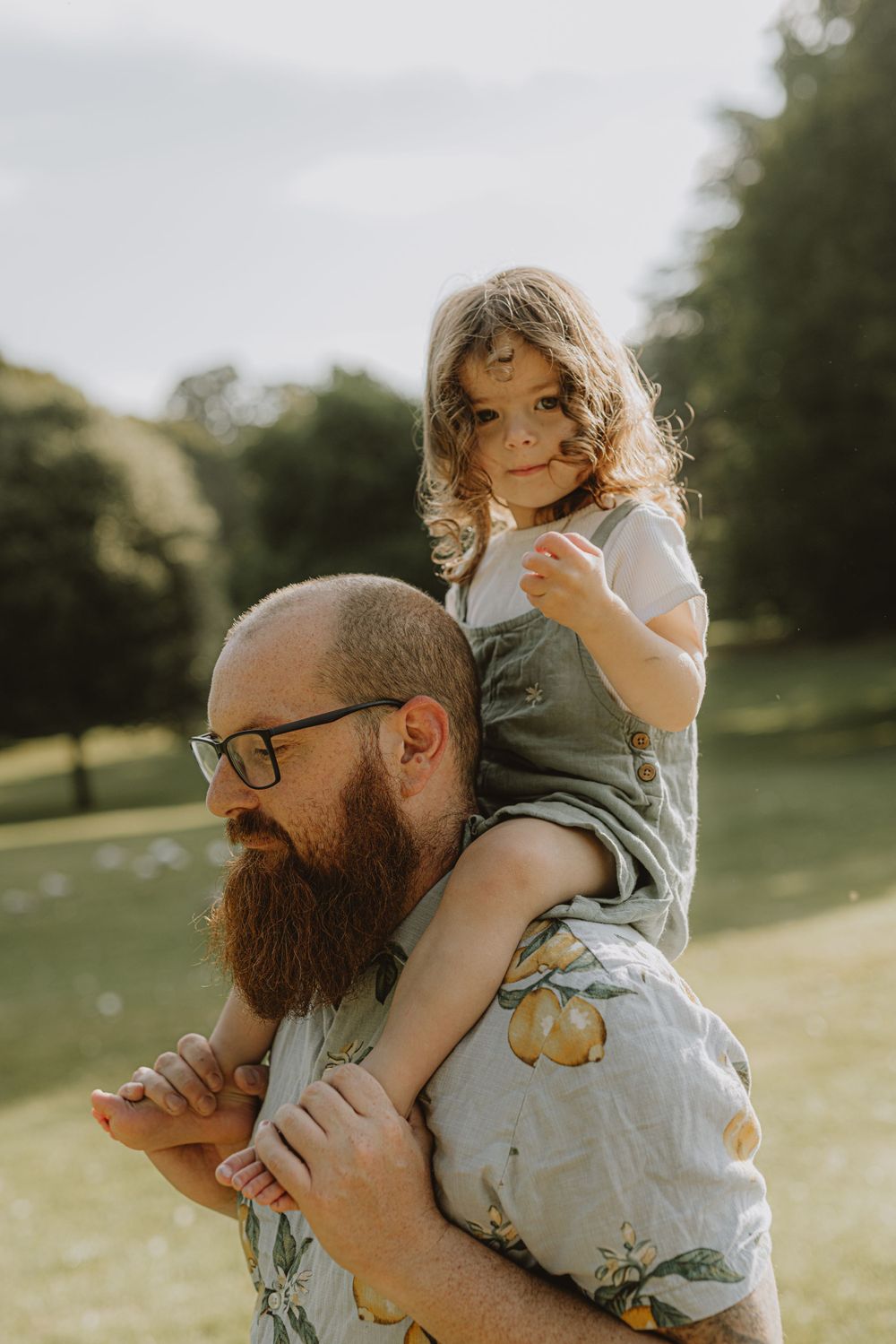 A bearded man in glasses wearing a floral shirt carries a child on his shoulders in a sunny outdoor setting.