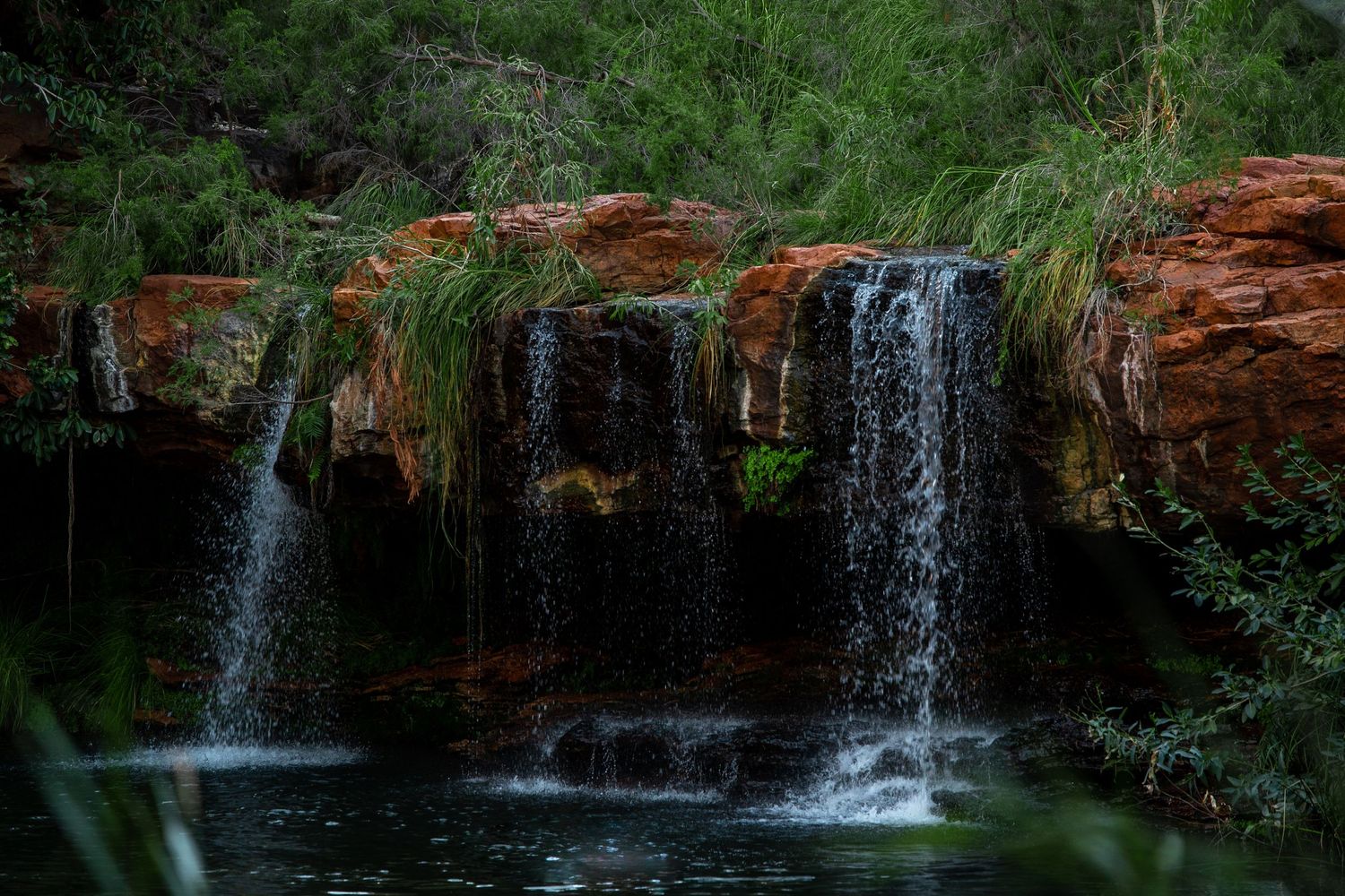 Multiple cascading waterfalls flowing over red rock formations into a serene pool surrounded by lush vegetation.