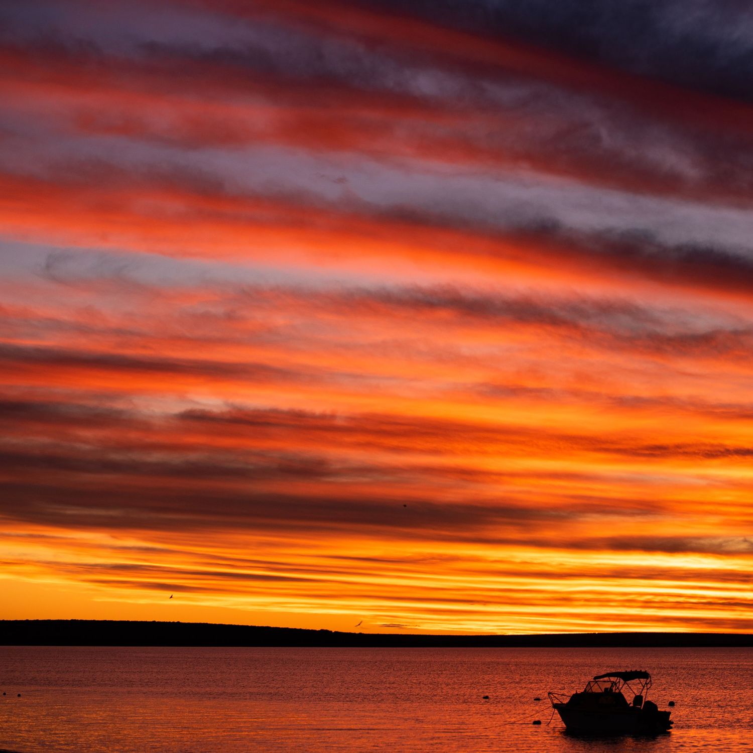 A silhouetted boat on calm waters during a vibrant orange and red sunset with dramatic striped clouds.