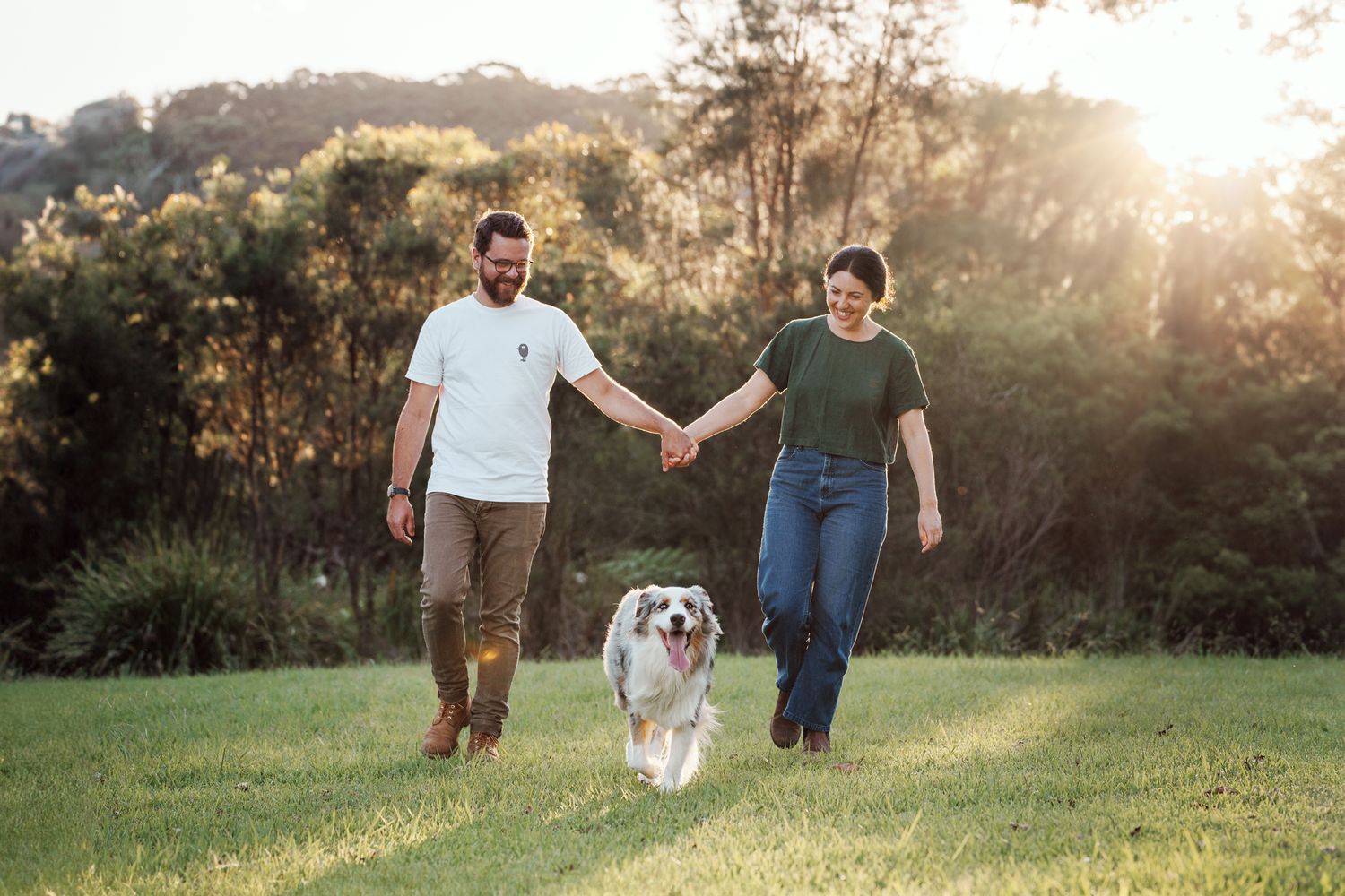 a couple walking hand in hand towards the camera during golden hour and their dog is walking in between them