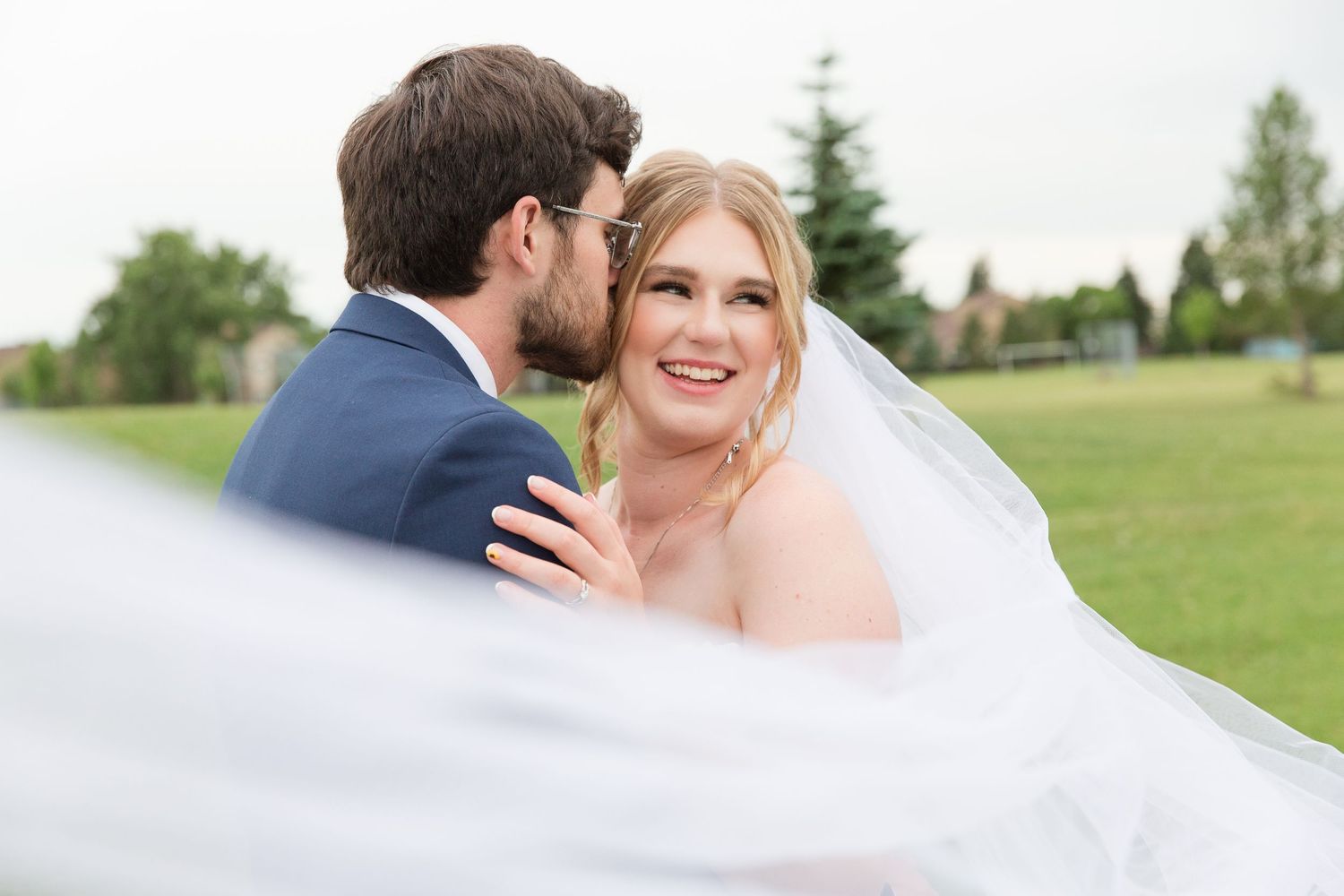 A romantic wedding moment captured outdoors on a lush green lawn with flowing veil in the foreground.