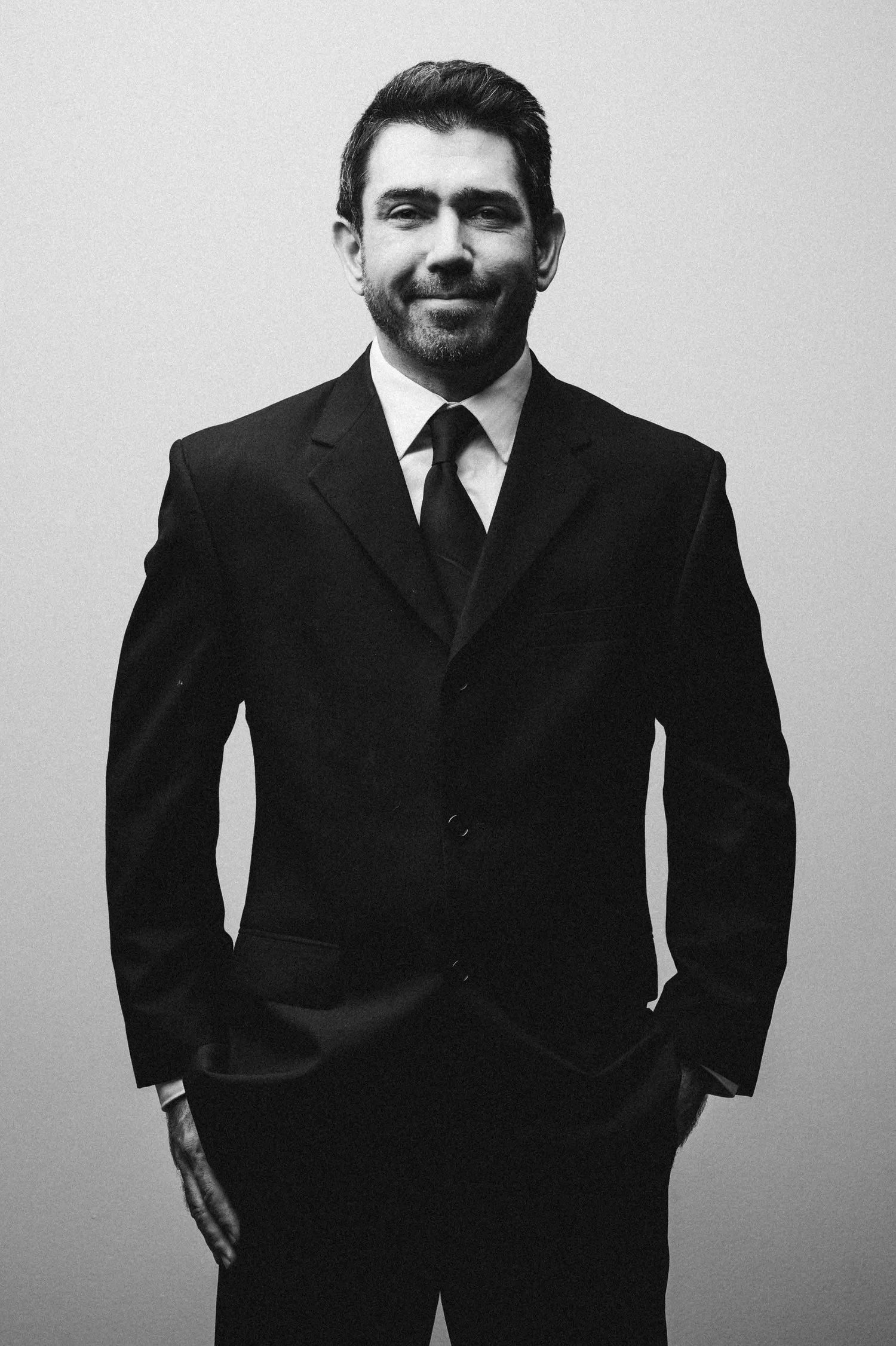 A black and white portrait of a person in a dark suit and tie smiling at the camera.