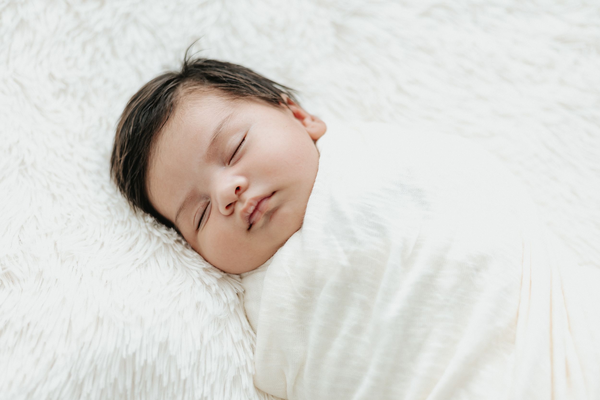 Sleeping baby wrapped in white blanket against soft white background.