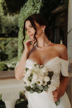 Elegant bride in off-shoulder white gown holds a classic white rose and eucalyptus bouquet in outdoor setting.