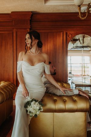 A bride in an elegant white dress poses gracefully against a vintage leather couch in a wood-paneled room.